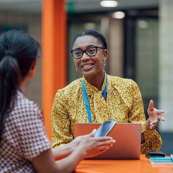 Two female teachers sitting at a table, having a discussion in the lobby area in the school they work at in Gateshead, North East England. They are talking about school issues together while using a laptop.