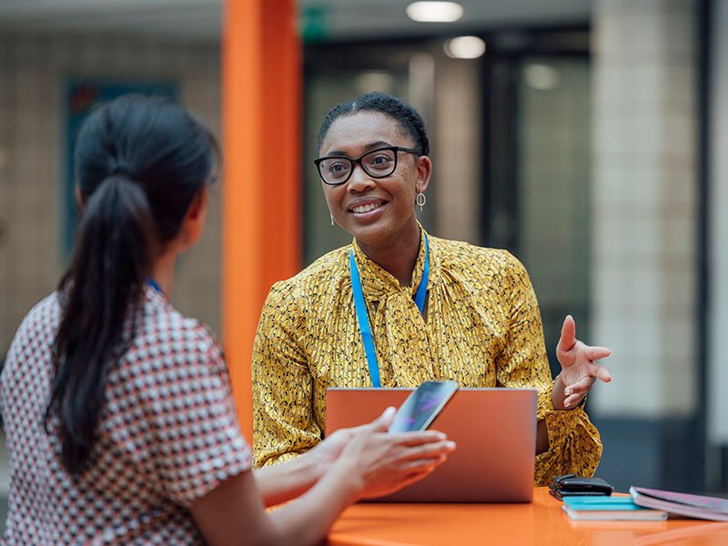 Two female teachers sitting at a table, having a discussion in the lobby area in the school they work at in Gateshead, North East England. They are talking about school issues together while using a laptop.